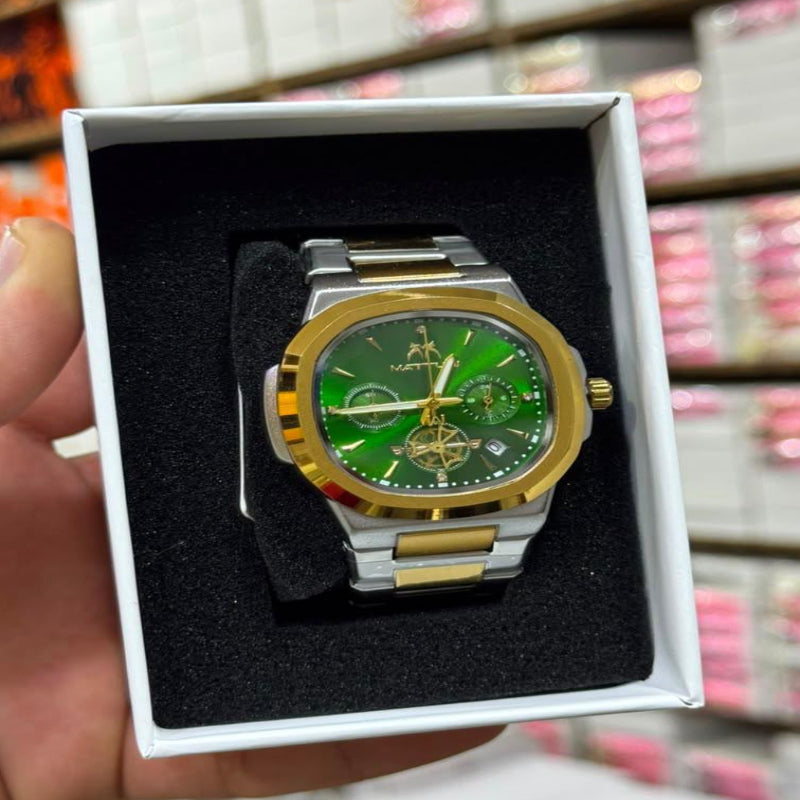 A gold and silver-tone wristwatch with a green dial, presented in a white box held by a hand. The background shows a blurred retail store setting with shelves of boxes.