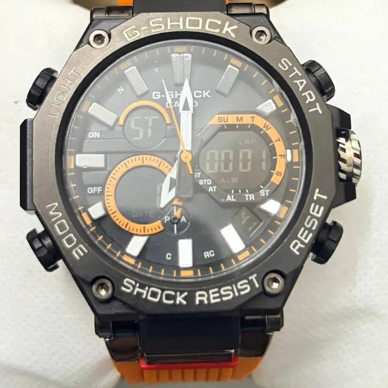 A close-up of a G-Shock analog-digital wristwatch with a black and gray case, orange accents on the dial and strap, and black rubber strap ends with orange tabs, photographed against a white textured background.