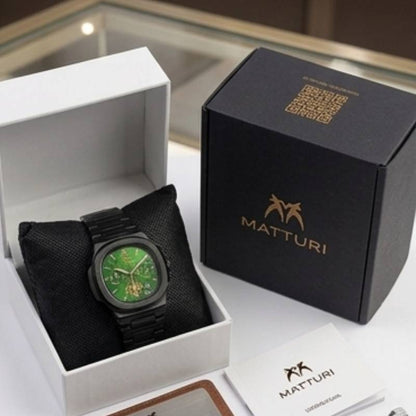 A matte black Matturi watch with a green dial and gold details, displayed inside an open white presentation box next to its black branded packaging box.