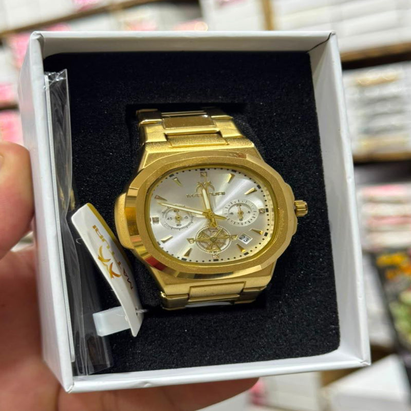 A gold-tone stainless steel watch with a white dial and three sub-dials, presented inside a white gift box with black foam insert. A hand is holding the box on the left side, with a blurred retail store background.