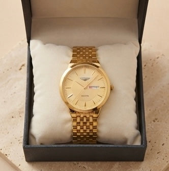 A gold-tone wristwatch with a matching metal link bracelet and a cream-colored dial, presented inside an open black gift box with a cream cushion.