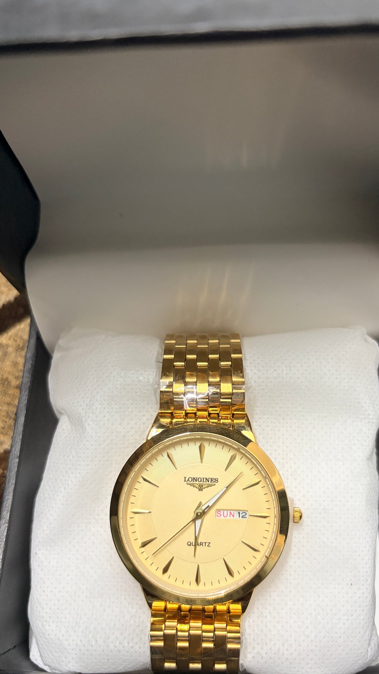 A gold-tone wristwatch with a cream-colored dial and matching gold link bracelet, presented inside an open gray gift box with white cushioning.