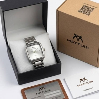 A silver-tone Matturi wristwatch with a square silver dial and metal link bracelet, displayed inside an open black presentation box with a white cushion. A closed kraft paper Matturi box, a card, and a manual are arranged beside it on a clean white background.