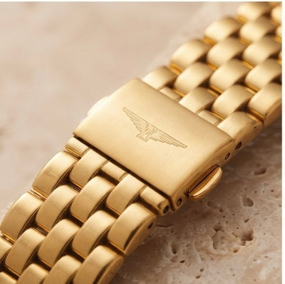 Close-up of a gold-tone metal link watch band with a rectangular clasp engraved with a winged logo, resting on a light beige textured surface.