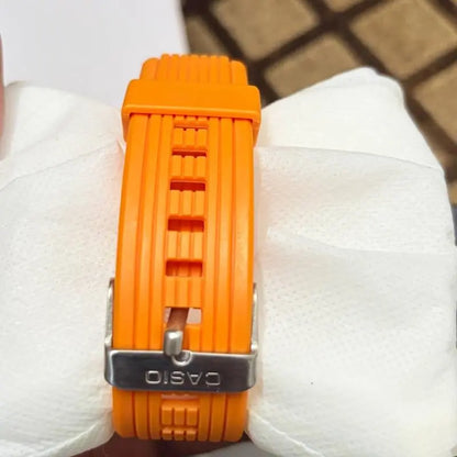 Bright orange silicone watch strap with a silver metal buckle engraved with CASIO, resting on white textured paper.