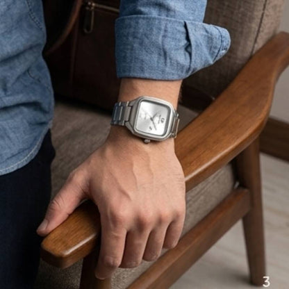 A silver-toned square analog watch with a white dial and metal link bracelet worn on a person's wrist, shown in a real indoor setting with a wooden chair and upholstered seat visible.