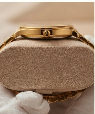 Gold-tone watch band and case resting on a beige textured watch cushion, with a white gloved hand partially visible at the bottom.